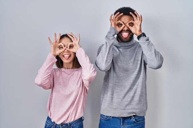 Jovem casal hispânico juntos fazendo gesto ok como binóculos enfiando a língua para fora os olhos olhando por entre os dedos expressão louca