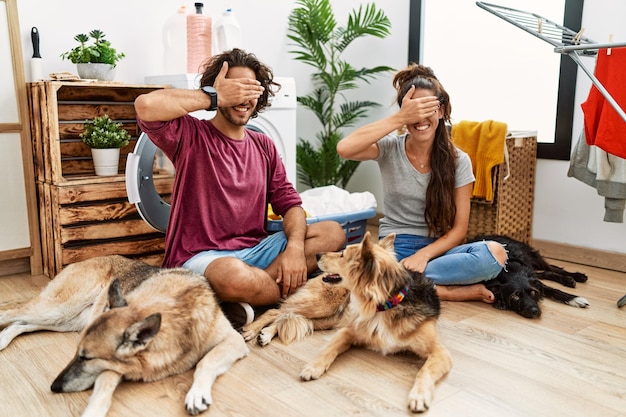 Jovem casal hispânico lavando roupa com cachorros sorrindo e rindo com a mão no rosto cobrindo os olhos de surpresa. conceito cego.