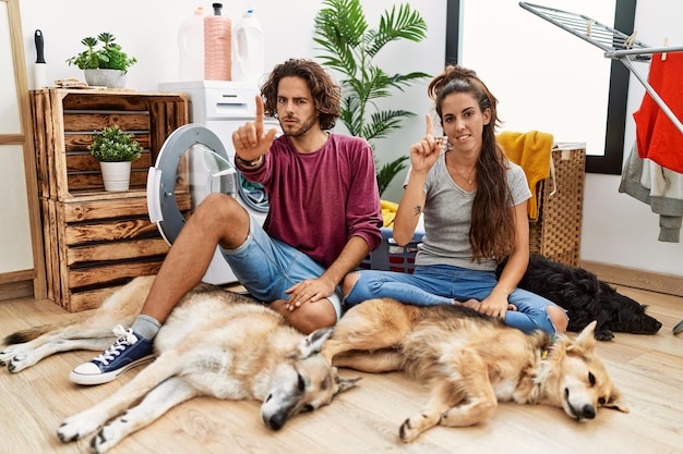 Jovem casal hispânico lavando roupa com cães apontando com o dedo para cima e expressão de raiva, não mostrando nenhum gesto