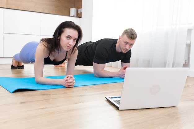 Jovem casal homem e mulher fazendo exercícios online em casa usando laptop, fazendo esportes online com o treinador no chão em casa