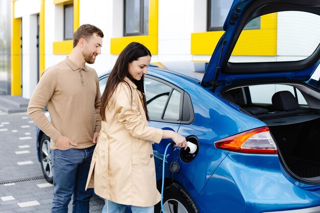 Jovem casal homem e mulher viajando de carro elétrico parando na estação de carregamento Namorada segurando o carregador olhando para o carro e sorrindo
