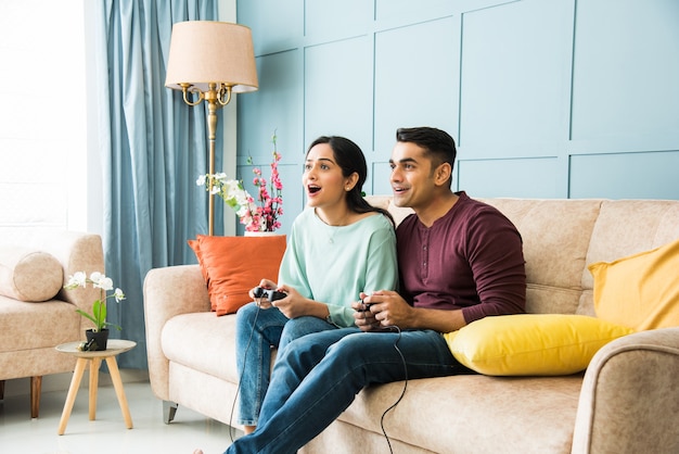 Jovem casal indiano asiático jogando videogame usando joystick ou controlador enquanto está sentado no sofá ou sofá