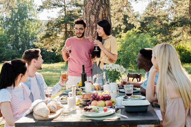 Jovem casal intercultural feliz com taças de vinho tinto anunciando seu noivado para amigos reunidos à mesa para jantar ao ar livre