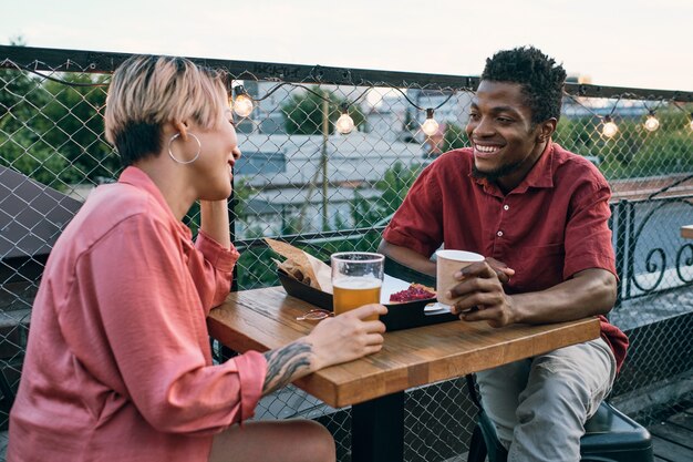 Jovem casal intercultural feliz tomando drinques e lanches em um café ao ar livre