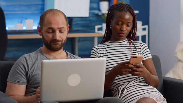 Jovem casal interracial usando tecnologia na sala de estar. Marido caucasiano trabalhando em um laptop moderno e uma esposa afro-americana olhando para o smartphone enquanto relaxam juntos no sofá