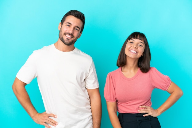 Jovem casal isolado em fundo azul posando com os braços no quadril e sorrindo