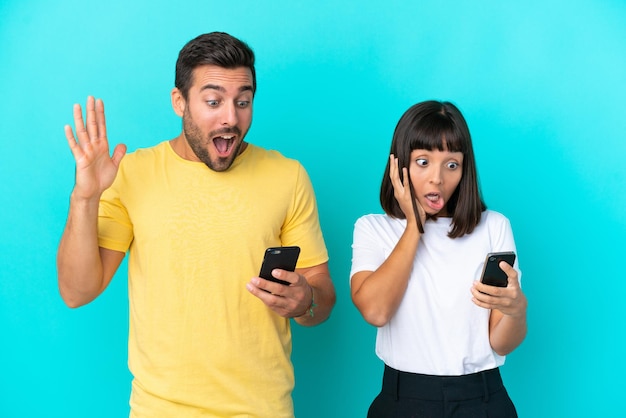 Foto jovem casal isolado em um fundo azul, olhando para a câmera enquanto usa o celular com expressão de surpresa