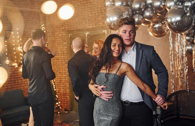 Foto jovem casal juntos de pé contra seus amigos na sala decorada de natal e comemorando o ano novo.