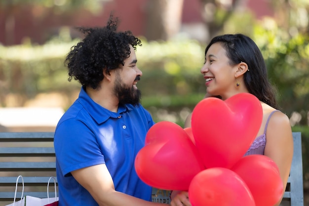 Jovem casal latino mexicano rindo e conversando no dia dos namorados com balões vermelhos em forma de coração
