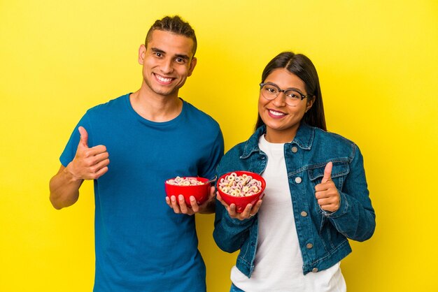 Foto jovem casal latino segurando uma tigela de cereais isolada em fundo amarelo sorrindo e levantando o polegar