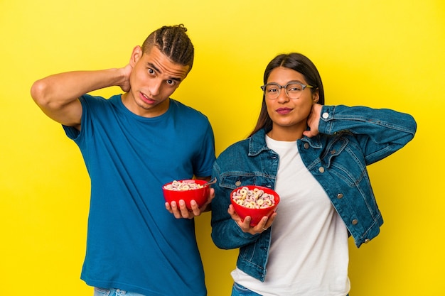 Foto jovem casal latino segurando uma tigela de cereais isolada na parede amarela tocando a parte de trás da cabeça, pensando e fazendo uma escolha.