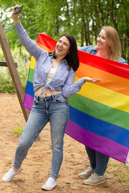 Jovem casal LGBT de lésbicas sorri enquanto segura a bandeira do Orgulho LGBTQ do arco-íris selfie