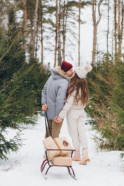 Jovem casal lindo abraços e beijos em uma floresta de coníferas de inverno e carrega caixas com presentes em um trenó. um parque com árvores de natal ao fundo. espírito de natal. tingimento.