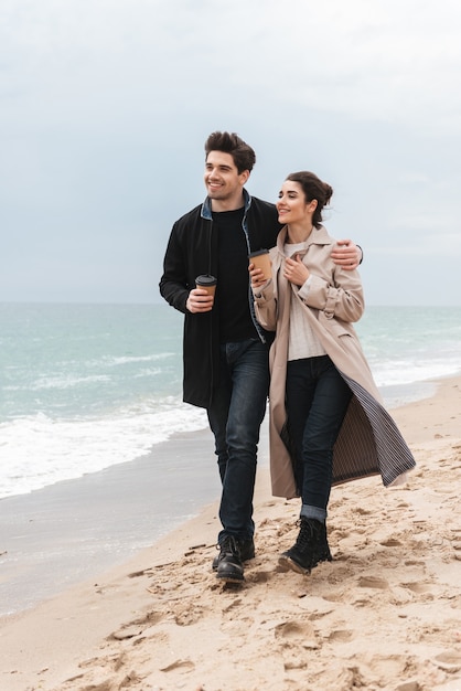 Jovem casal lindo de casacos caminhando na praia, tomando café para viagem