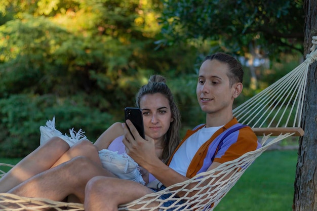 Jovem casal moderno sentado em uma rede em feriados e férias enquanto olha para o smartphone cercado pela natureza