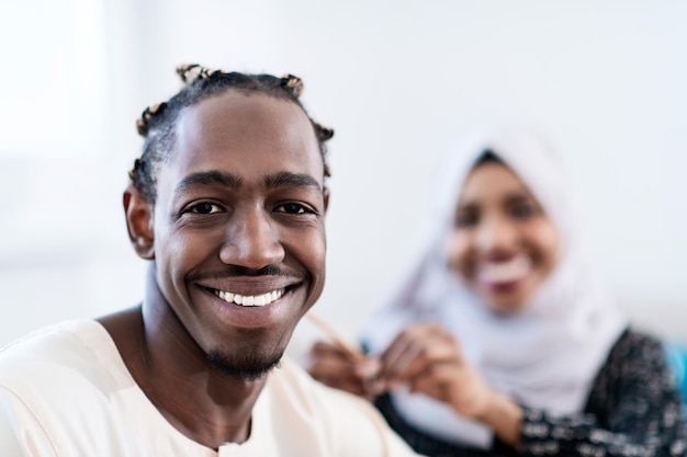 jovem casal muçulmano afro moderno tendo momentos românticos em casa enquanto mulher fazendo penteado para marido feminino vestindo roupas tradicionais de hijab islâmico do sudão