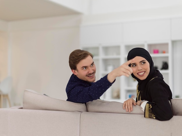 Jovem casal muçulmano vestindo roupas de hijab islâmicos sentados no sofá assistindo TV juntos durante o mês do Ramadã em uma casa moderna.