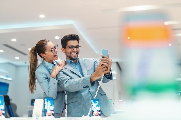 Jovem casal multicultural atraente vestido elegante tomando selfie com novo telefone inteligente na loja de tecnologia.