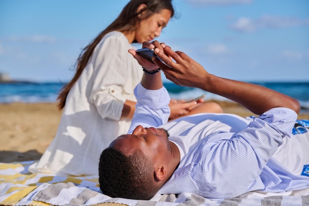 Jovem casal multicultural deitado na praia usando smartphones