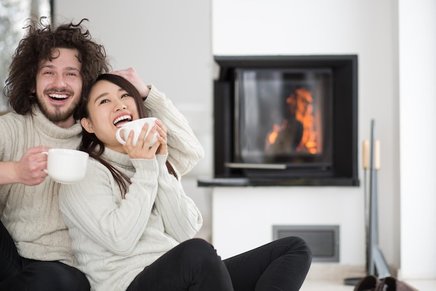 Jovem casal multiétnico romântico sentado no chão em frente à lareira em casa, conversando e tomando café no dia frio de inverno