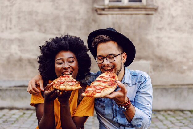 Jovem casal multirracial fofo sentado ao ar livre e comendo pizza.