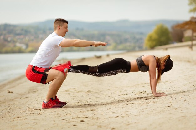 Jovem casal musculoso tendo treino juntos na praia. Garoto bonito fazendo exercícios de agachamento, sua namorada fofa fazendo flexão.