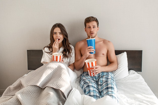 Jovem casal na cama. Um intrigado homem e mulher bonita estão comendo pipoca e assistindo TV juntos no quarto