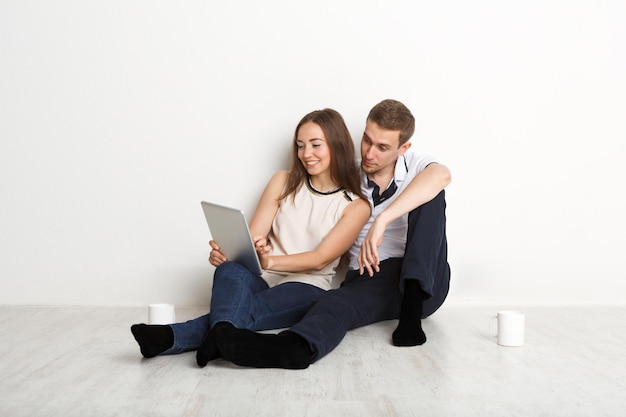 Jovem casal navegando na web em um laptop sentado no chão da sala vazia de um apartamento novo no fundo branco, copie o espaço