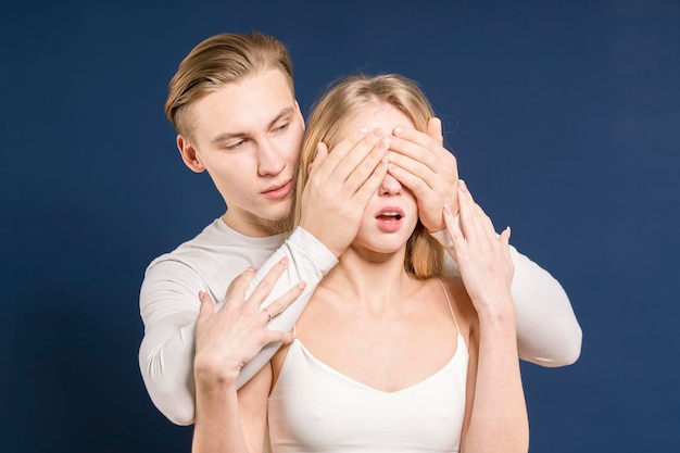 Jovem casal no estúdio O cara cobre as mãos com os olhos de uma garota Fundo azul escuro