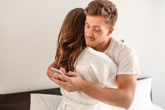 Jovem casal no quarto. sorridente homem infiel está traindo e mandando mensagens por telefone enquanto abraça a namorada