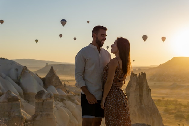 Jovem casal olhando nos olhos um do outro em primeiro plano assistindo os típicos balões de ar quente da área da capadócia voando ao nascer do sol