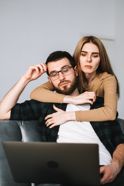 Jovem casal pensativo usando laptop e descansando juntos no sofá em casa.