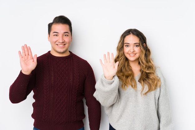 Jovem casal posando em uma parede branca sorrindo alegre mostrando número cinco com os dedos.