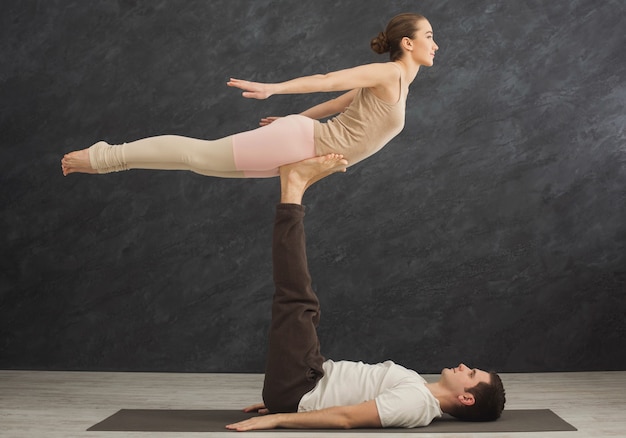 Jovem casal praticando acroyoga na esteira na academia juntos. Mulher voando, cópia espaço, vista lateral. Ioga de parceiro, flexibilidade, conceito de confiança