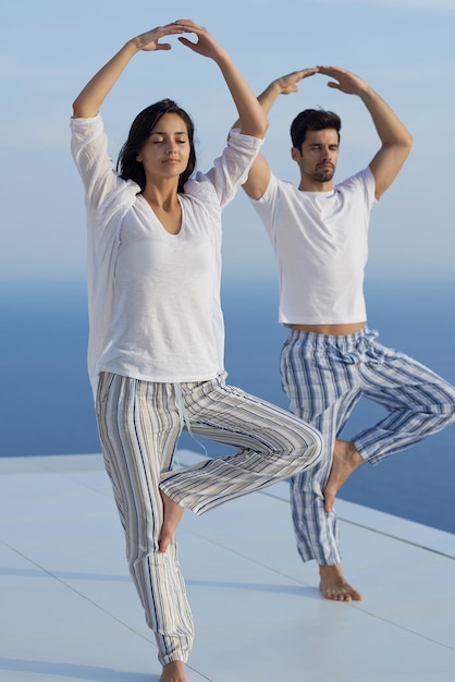 jovem casal praticando ioga ao pôr do sol no terraço doméstico moderno com oceano e pôr do sol ao fundo