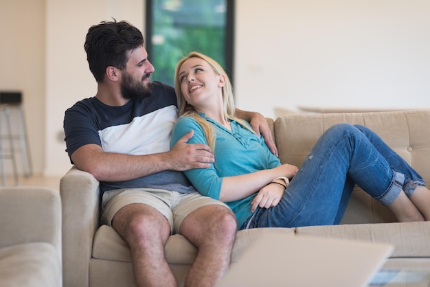 Jovem casal relaxa no sofá na sala de estar de luxo, usando um laptop e controle remoto