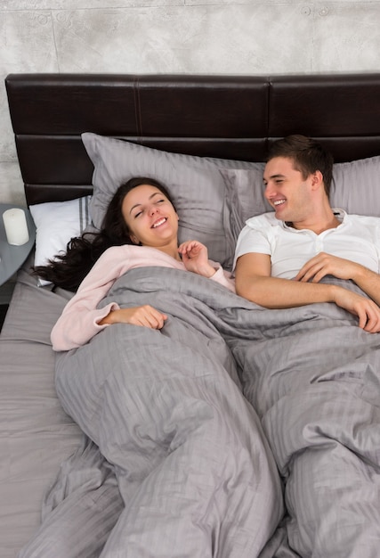 Jovem casal rindo e de pijama enquanto estava deitado na cama no quarto em estilo loft com cores cinza