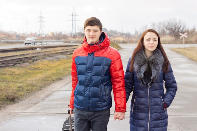 Jovem casal romântico carregando uma mala enquanto caminha por uma estrada rural de mãos dadas enquanto partem para as férias ou lua de mel