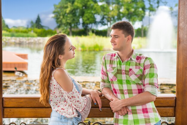 Jovem casal romântico encostado na grade do gazebo de madeira e olhando um para o outro em frente a uma cachoeira artificial em um parque com lago, fonte e árvores diferentes