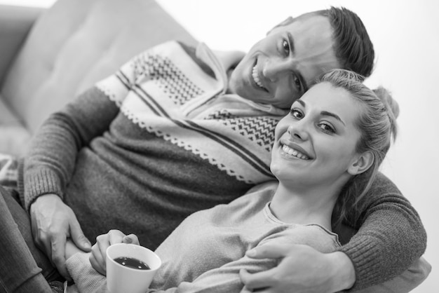 Jovem casal romântico sentado no sofá em frente à lareira em casa, olhando um para o outro, conversando e bebendo café dia de outono