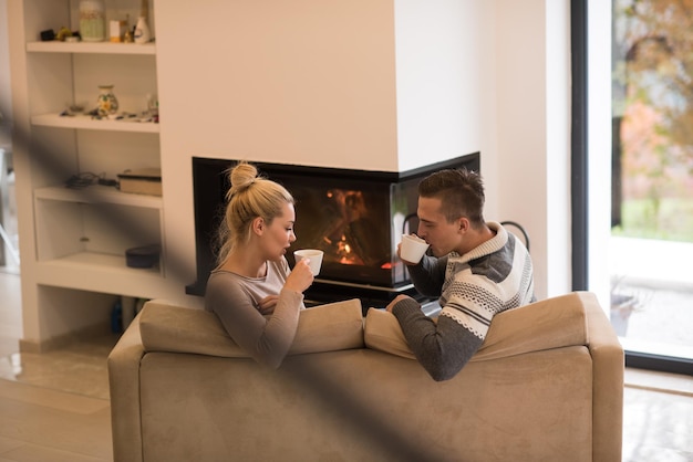 Foto jovem casal romântico sentado no sofá em frente à lareira em casa, olhando um para o outro, conversando e tomando café no dia de outono