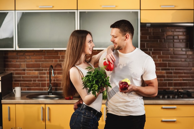 Jovem casal saudável e nutrição adequada. homem e mulher posando na cozinha segurando verduras e legumes. jovens se entreolham