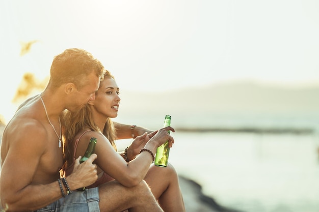 Jovem casal se divertindo e bebendo cerveja na praia à beira-mar. Desfrutando de seu amor.