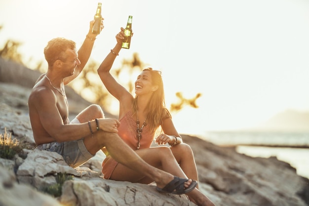 Jovem casal se divertindo e bebendo cerveja na praia à beira-mar. Desfrutando no belo pôr do sol e seu amor.