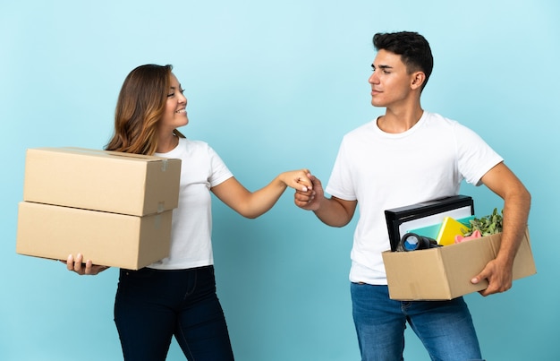 Jovem casal se mudando para a nova casa entre caixas em um aperto de mão azul após um bom negócio