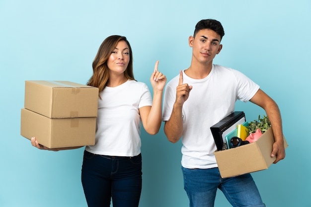 Jovem casal se mudando para uma nova casa entre caixas em azul, mostrando e levantando um dedo em sinal dos melhores