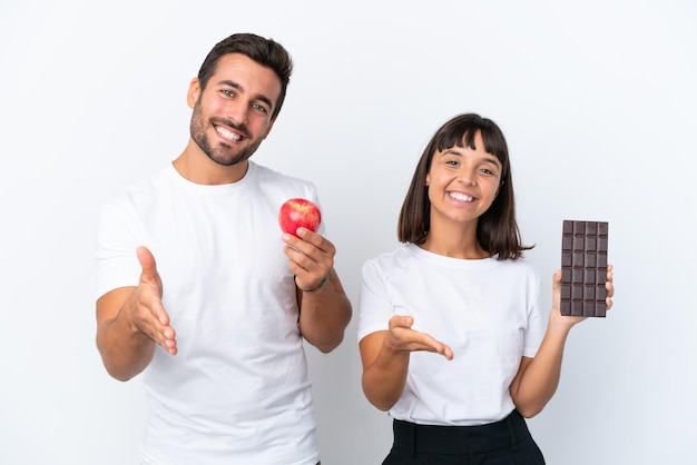 Jovem casal segurando chocolate e maçã isolado no fundo branco, apertando as mãos para fechar um bom negócio