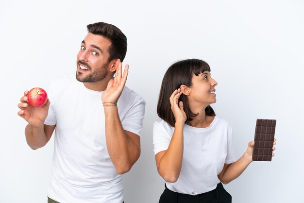 Jovem casal segurando chocolate e maçã isolado no fundo branco ouvindo algo colocando a mão na orelha