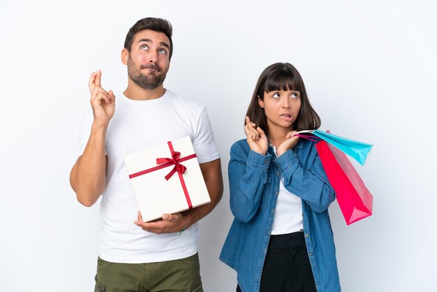 Jovem casal segurando sacolas de compras e presentes isolados no fundo branco com dedos cruzando e desejando o melhor