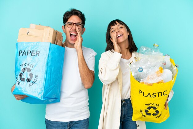 Jovem casal segurando um saco cheio de plástico e papel para reciclar isolado em fundo azul gritando e anunciando algo